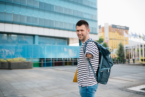 Portrait of a happy young adult male university student with an amputated arm standing in front of the university building, looking at the camera