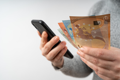 Young woman with a gray pullover holds mobile phone and euro money in her hands
