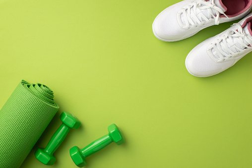 Sports concept. Top view photo of white shoes green yoga mat and dumbbells on isolated green background with empty space