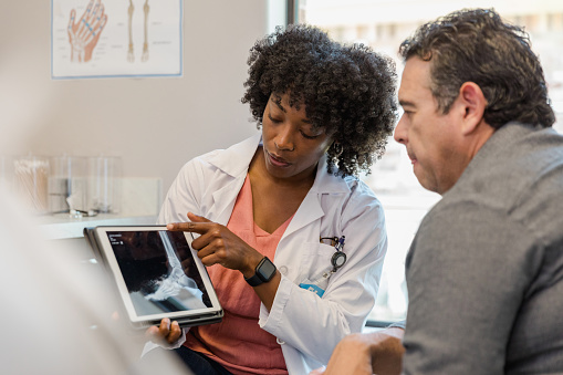 The mid adult female orthopedic surgeon points to the x-ray of the patient's foot on digital tablet as she explains treatment options to the mature adult man.