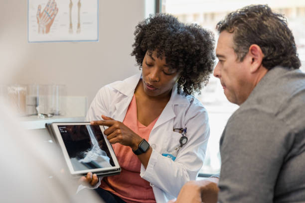 Female orthopedic surgeon points to foot x-ray on digital tablet The mid adult female orthopedic surgeon points to the x-ray of the patient's foot on digital tablet as she explains treatment options to the mature adult man. orthopedist stock pictures, royalty-free photos & images