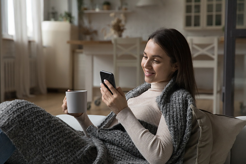 Happy millennial gen Z smartphone user girl wrapped in warm knitted plaid, drinking hot tea, coffee, cocoa, using online app on smartphone, relaxing on couch at cozy home