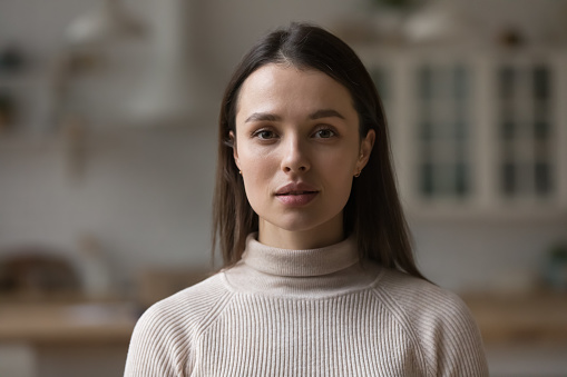 Positive serious millennial model girl home female head shot portrait. Beautiful young adult Caucasian woman looking at camera, posing in apartment. Front profile picture