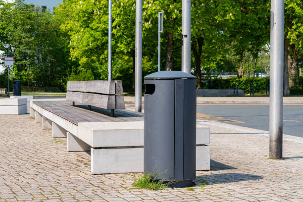 A metal trash bin and a long bench in a city park.