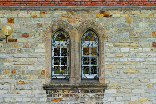 Stone wall of an old building with two arched windows. Windows are closed with decorate metal grid. Lantern on wall.