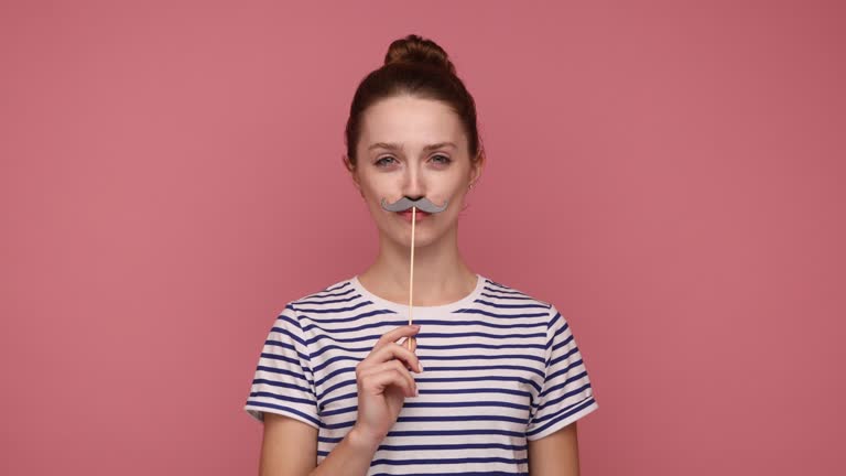 Woman keeping fake paper mustache and looking at camera with happy expression, having festive mood.