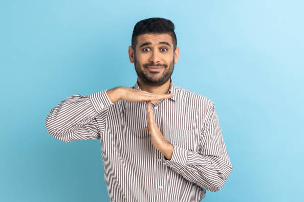 Handsome businessman standing with serious face and looking at camera with timeout gesture. Timeout. Portrait of young handsome businessman standing with serious face and looking at camera with timeout gesture, wearing striped shirt. Indoor studio shot isolated on blue background. time out signal stock pictures, royalty-free photos & images