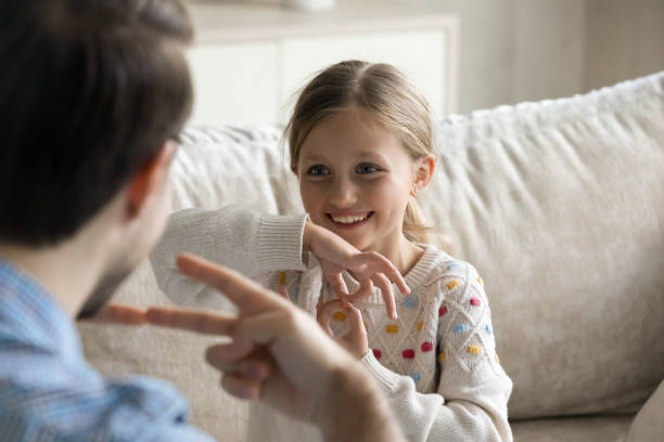 Happy small girl using sign language, communicating with father. Joyful cute little preteen child girl using sign language communicating with caring young father or professional male therapist, sitting together on sofa, making success in skills improvement. speak no evil stock pictures, royalty-free photos & images