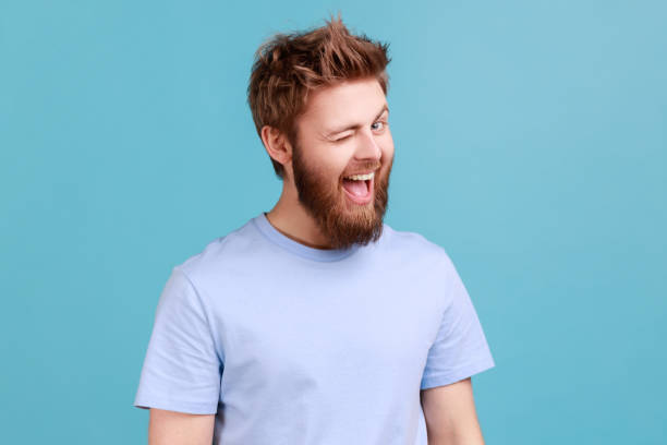 Man in blue T-shirt in good mood, smiling and winking at camera keeps mouth open flirting expression Portrait of bearded man being in good mood, smiling broadly and winking at camera, keeps mouth open, having flirting facial expression. Indoor studio shot isolated on blue background. winking stock pictures, royalty-free photos & images