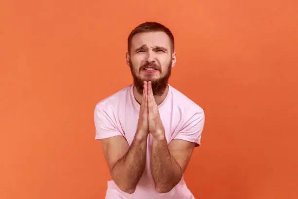 Portrait of bearded man folding hands in prayer, closing eyes and talking to god, asking for help, expressing gratitude, wearing pink T-shirt. Indoor studio shot isolated on orange background.