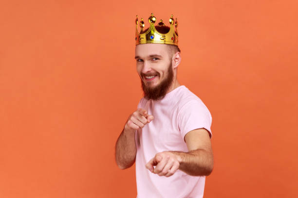 istock Man looking smiling at camera, posing with crown on head, pointing at you with happy expression. 1405695957