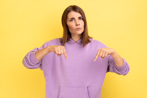 Portrait of serious strict confident bossy woman pointing fingers down, asking to act here and now immediately, wearing purple hoodie. Indoor studio shot isolated on yellow background.