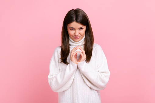 Tricky cunning pretty female standing looks at camera, planning something cunful, pranking somebody, wearing white casual style sweater. Indoor studio shot isolated on pink background.