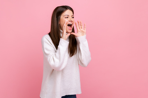 Side view portrait of brunette female standing, holding arms near wide open mouth and screaming, wearing white casual style sweater. Indoor studio shot isolated on pink background.