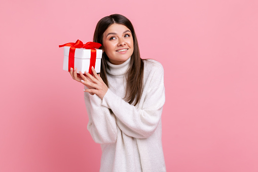 Good looking girl with dark hair holding wrapped present box, being interested what inside, smiling, wearing white casual style sweater. Indoor studio shot isolated on pink background.