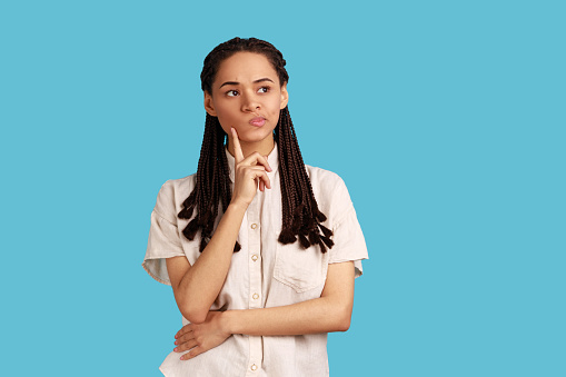 Portrait of thoughtful woman with black dreadlocks thinking about future, holding chin, having serious facial expression, wearing white shirt. Indoor studio shot isolated on blue background.