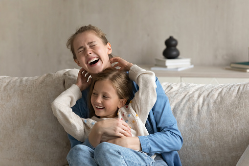 Overjoyed young mommy having fun with laughing little kid girl, joking playing together at home. Sincere happy two female generations family enjoying entertainment activity on weekend in living room.
