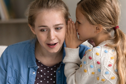 Curious happy young mother listening to secret, joyful preteen kid girl whispering on ear, telling confidential information or gossiping at home, trustful conversation, sincere relations concept.