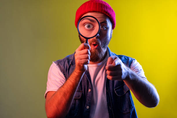 istock Man standing, holding magnifying glass, pointing to camera with surprised face. 1405696180