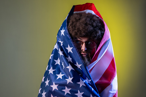 Portrait of hipster man with Afro hairstyle standing wrapped in american flag, looking away with funny facial expression and pout lips. Indoor studio shot isolated on colorful neon light background.