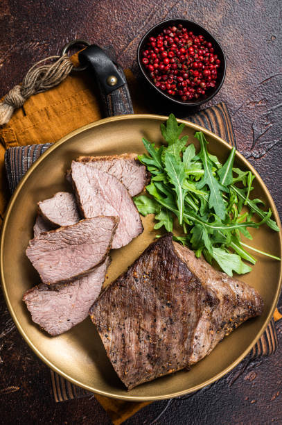istock Roast Tri Tip or sirloin bottom beef steak on a plate with arugula. Dark background. Top view 1405701312