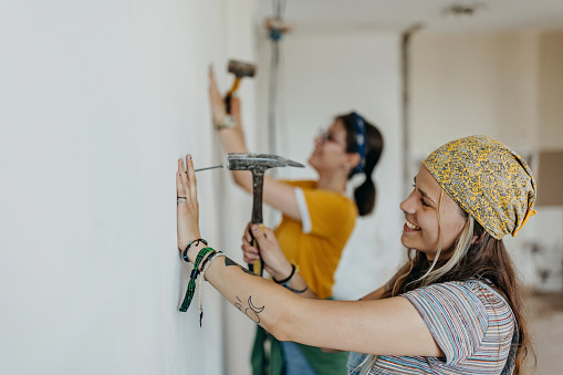 Two young Caucasian women are hammering nails into the wall for their home renovation project.