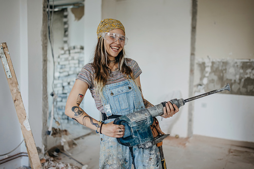 A young Caucasian woman is holding a pneumatic sledgehammer with a big smile on her face.