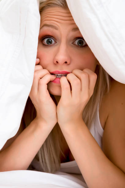 Terrified caucasian blond female laying underneath white blanket with hand covering mouth.