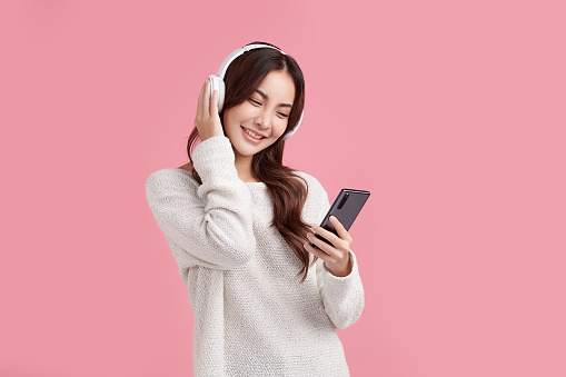 Happiness Beautiful Asian smiling young woman wearing technology wireless  headphones for listening the music via smart mobile phone on pink background,