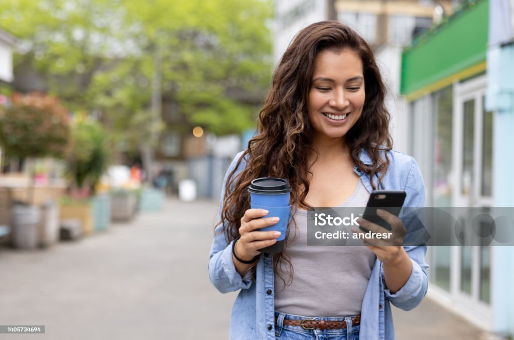 Happy woman drinking a cup of coffee on the move while texting on her phone Happy woman drinking a cup of coffee on the move while texting on her cell phone and smiling - lifestyle concepts Direction Stock Photo
