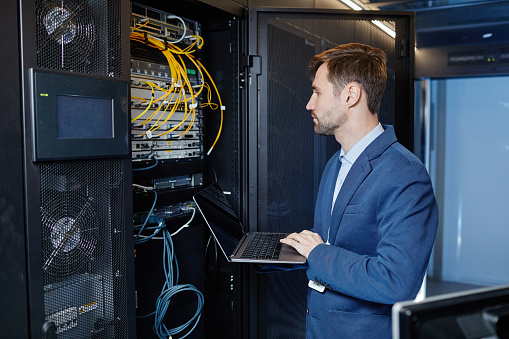 Side view portrait of Caucasian young man holding laptop while setting up internet network in server room