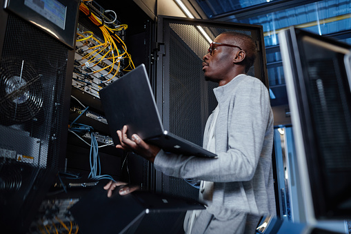 Side view portrait of black man working as IT engineer holding laptop while setting up internet network in server room