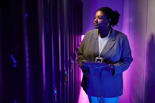 Portrait of female system administrator inspecting data network in server room lit by neon light, copy space