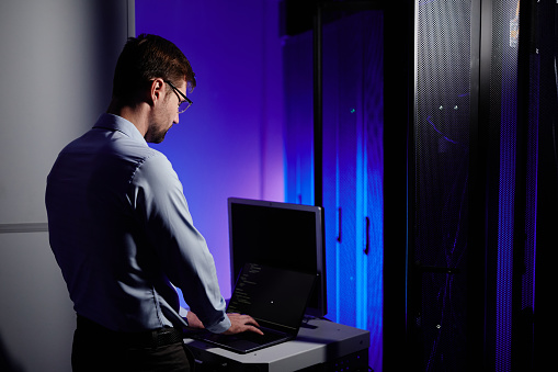 Back view of network engineer using laptop in dark server room lit by neon light, copy space