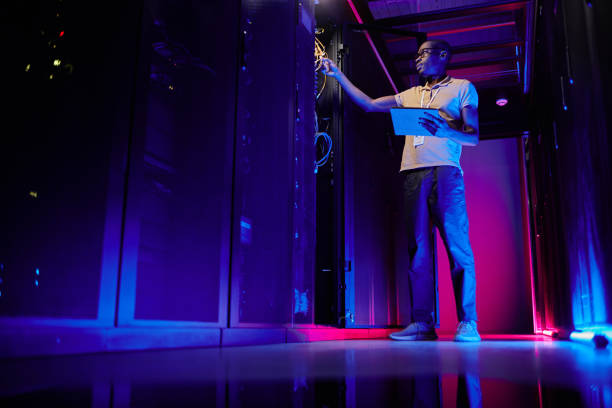 Technician in Neon Server Room Full length portrait of African American system administrator setting up server network in data center lit by neon light, copy space datacentre stock pictures, royalty-free photos & images