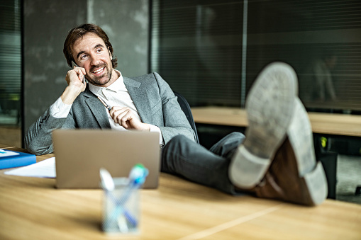 Happy businessman communicating over smart phone while resting his feet on the table in the office.