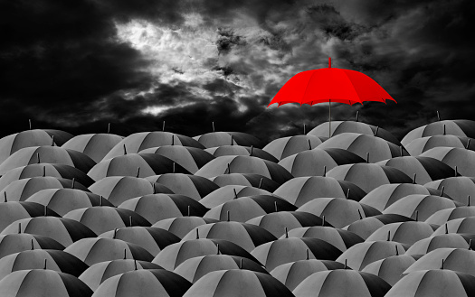 Red umbrella surrounded by many black umbrellas against dark cloudy sky with copy space.
Standing out from the crowd.