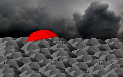 Red umbrella surrounded by many black umbrellas against dark cloudy sky with copy space.\nStanding out from the crowd.