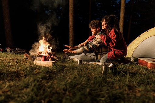 Young happy couple relaxing by the fire during camping night in nature. Copy space.