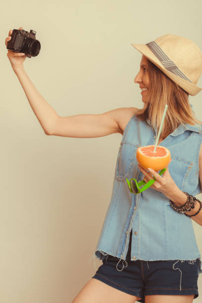 Happy tourist woman in straw hat taking selfie self picture with camera while holding grapefruit fruit in hand. Summer vacation holiday. Instagram filtered.
