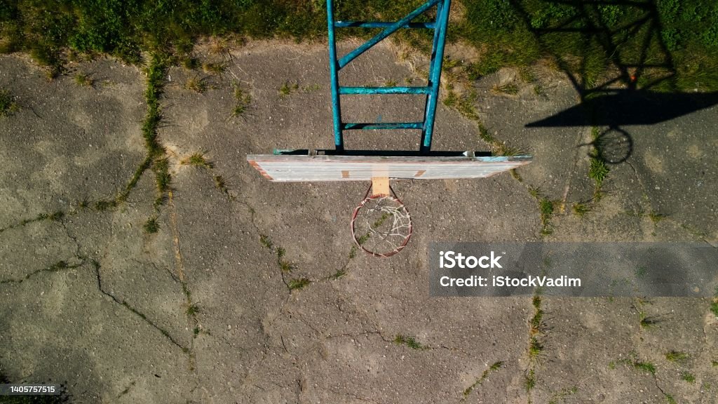 Old basketball backboard. Made from boards. Peeling paint and a battered basket. There is an old cracked asphalt on the site. Shot from above. Aerial photography. Basketball - Sport Stock Photo
