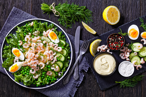 green salad of pink cocktail shrimps with lettuce, green peas, new potatoes, cucumber, dill and eggs on plate with mustard yogurt dressing and ingredients on wood table, horizontal view from above