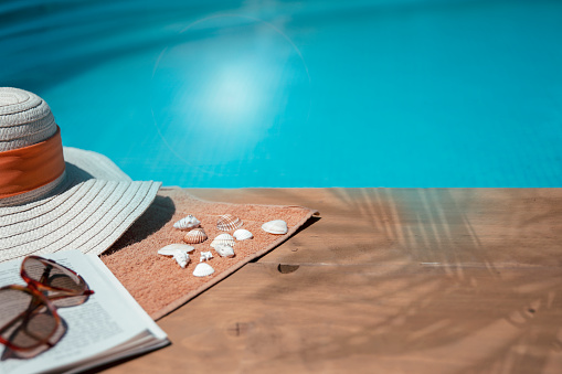 Sun hat, sunglasses, towel, shells and a book at the pool side in palm shade. Relaxing with bestseller book in summer holiday. Copy space