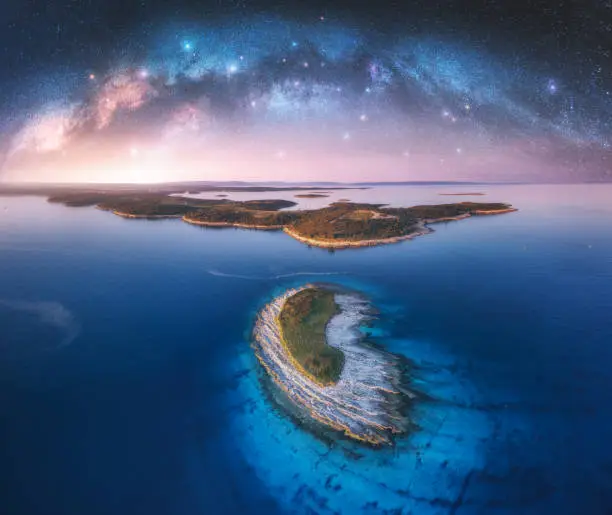 Photo of Milky Way arch and small island in the sea at summer night. Kamenjak cape, Adriatic sea, Croatia. Landscape with purple starry sky, arched milky way, sea coast, water, mountains. Top view. Panorama