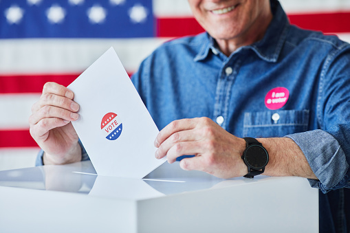 Close up of of smiling senior man putting ballot in bin against American flag in background, copy space