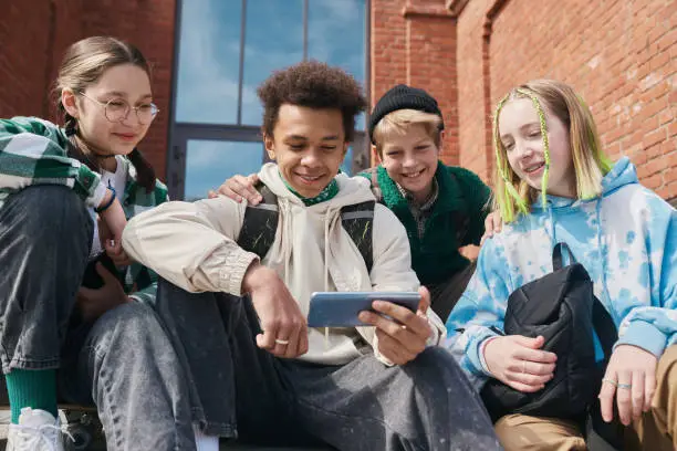 Group of positive school children watching video on smartphone and smiling while sitting outdoors