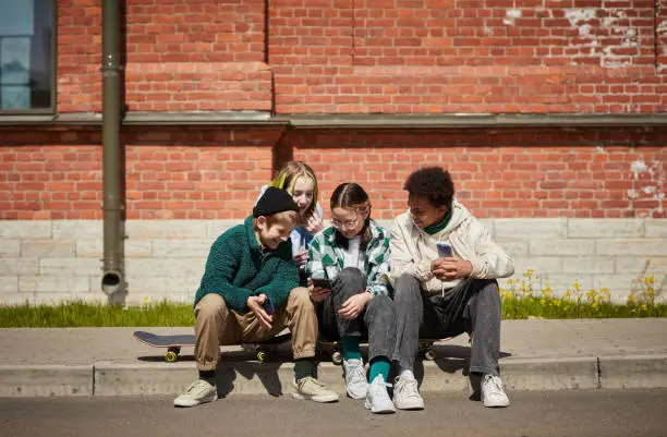 Group of teenagers sitting on skateboards outdoors and having fun of watching video online on their smartphones