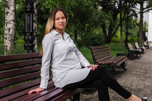 Pregnant young woman sitting on bench and holding hand on belly looking at camera outdoors.