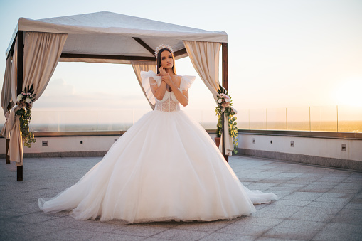 Beautiful bride posing outdoor at sunset in stunning dress and crown. Terrace and gorgeous bride making great enjoyment mood.