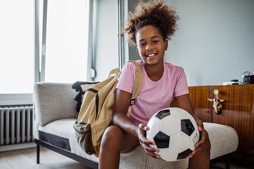 Adorable African-American teenage girl sitting on the sofa and smiling at the camera with a soccer ball in her hands and a backpack on her shoulder.